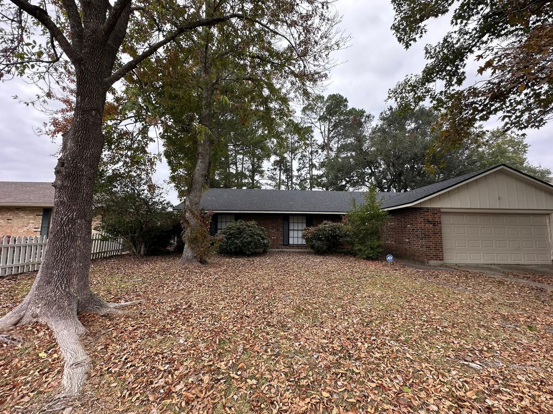 A single-story brick house with a front-facing garage, surrounded by large trees and a yard covered in fallen autumn leaves.