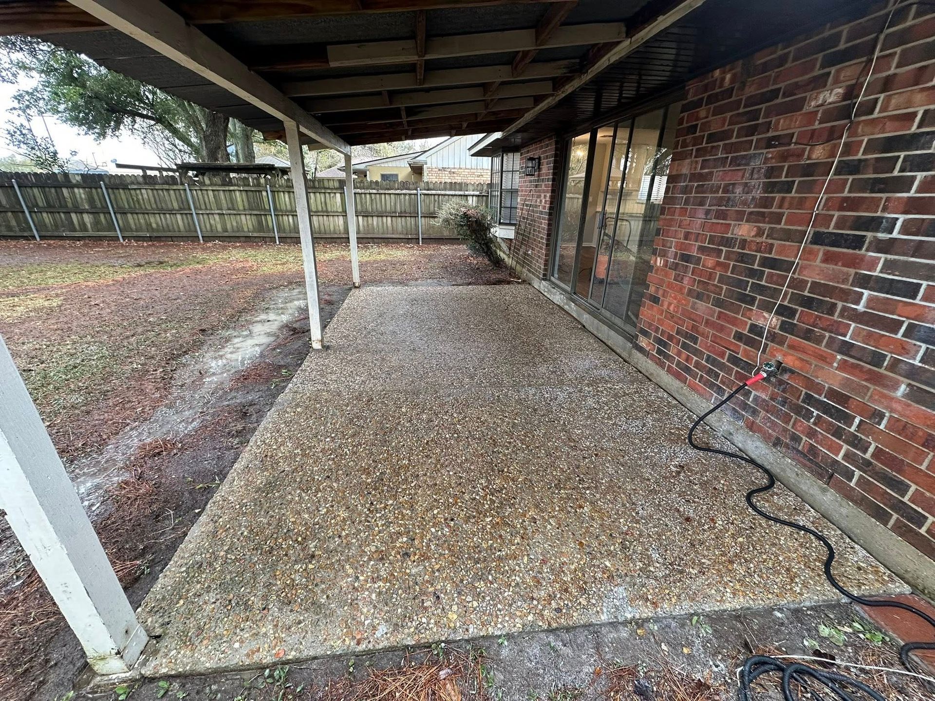 A covered patio with a pebble-dash concrete floor next to a red brick house wall, leading to a fenced backyard.