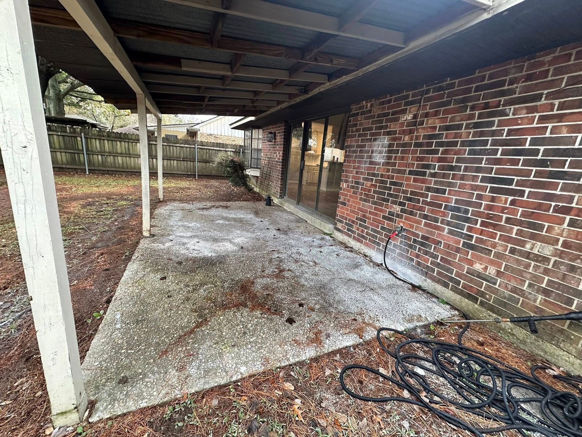 A concrete patio under a covered porch attached to a red brick house, with a coiled black hose in the foreground.