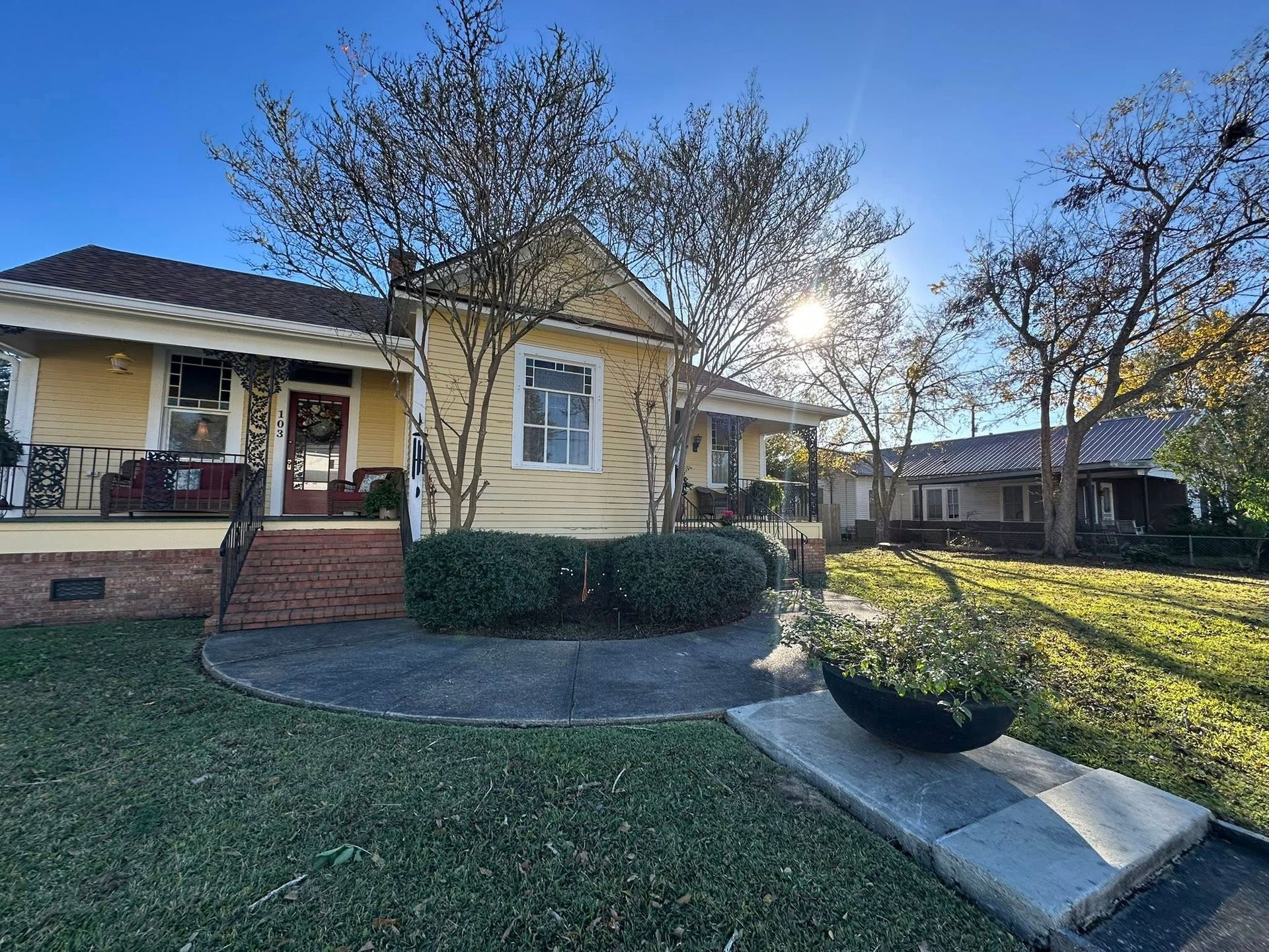 A sunny day view of a yellow house with a brick porch and steps, surrounded by green grass and trees.
