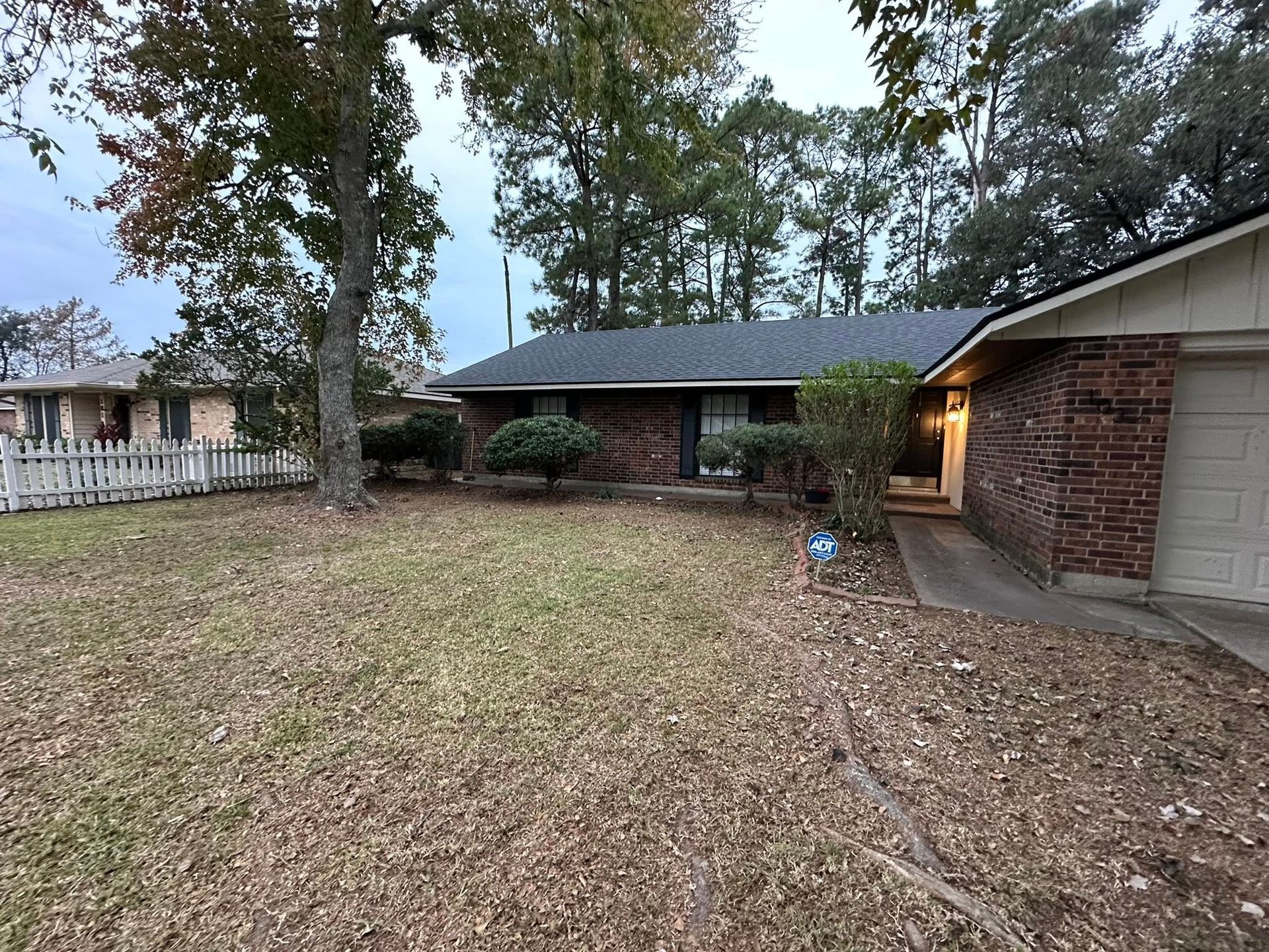 A brick house with a dark roof and large trees in the yard on an overcast day.