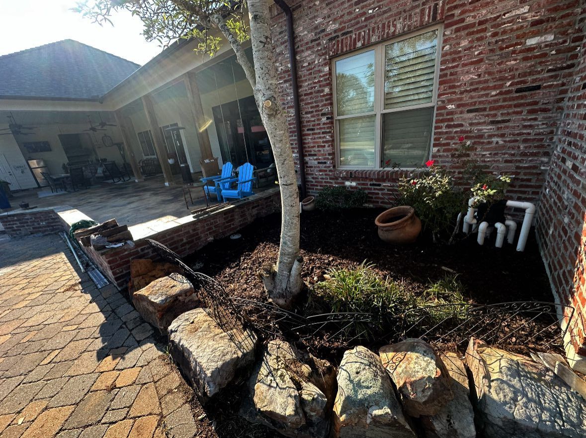 A brick house corner features a garden bed with a small tree, mulch, and large stone borders next to a patio.