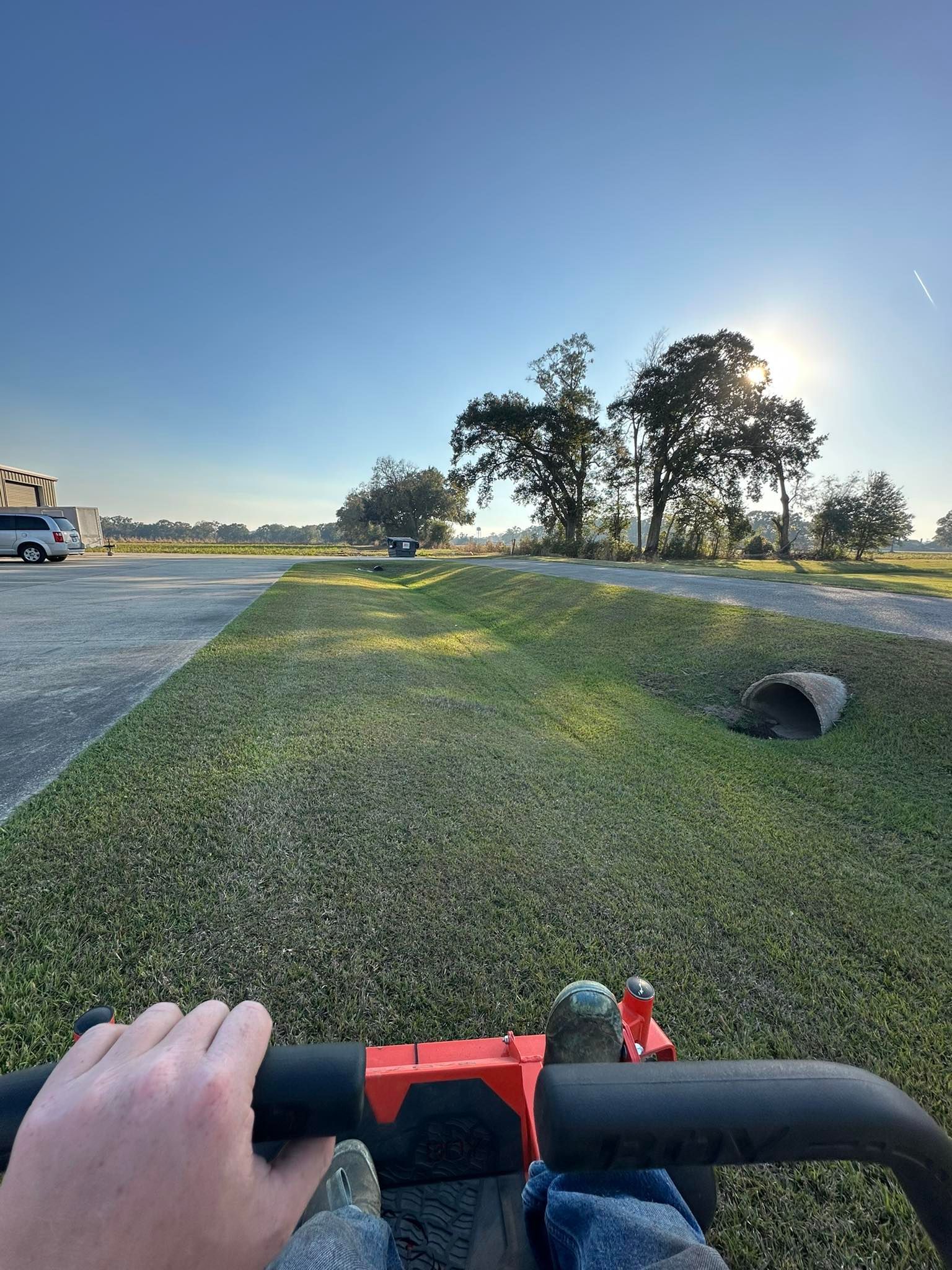 First-person view from a red lawn mower, showing a grassy ditch with a concrete culvert under a clear, sunny sky.