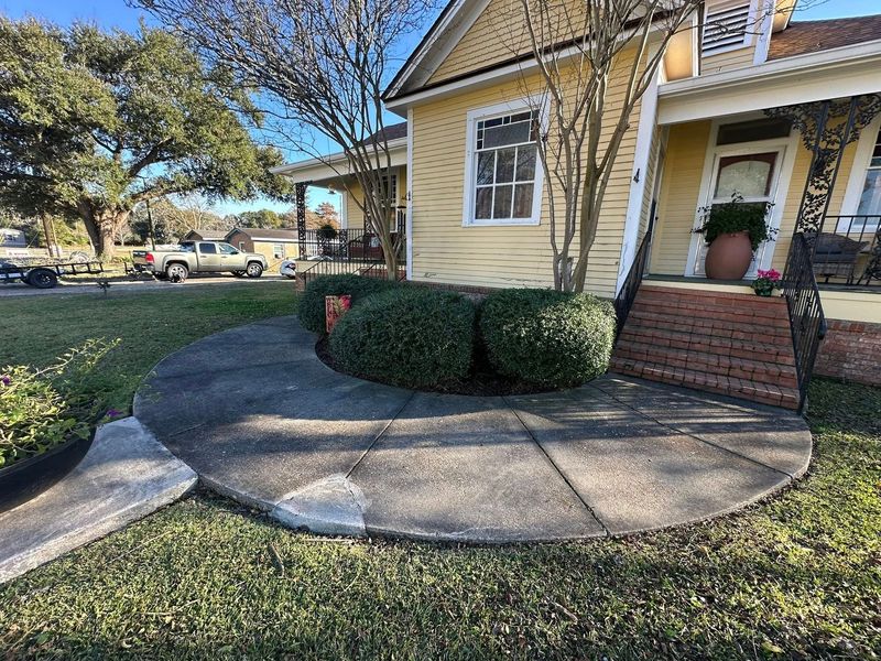 A yellow wooden house with a circular concrete patio in the front yard featuring three rounded green bushes.