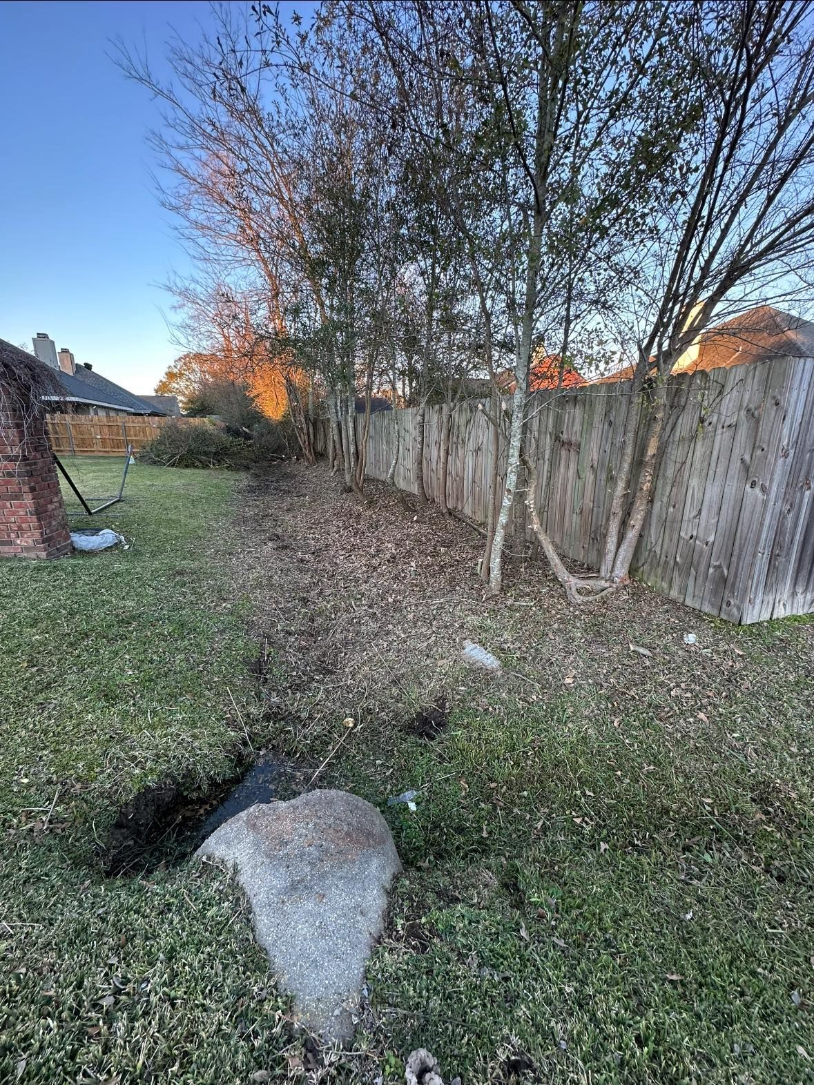 A backyard lawn with a large rock in the foreground, leading toward a wooden fence lined with trees under a blue sky.