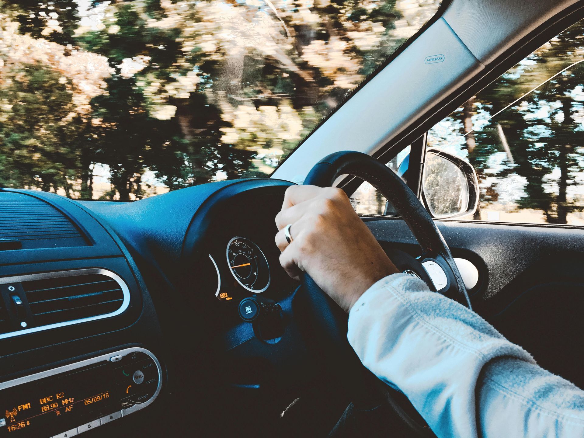 Person's hands on car steering wheel, driving on a road surrounded by trees.