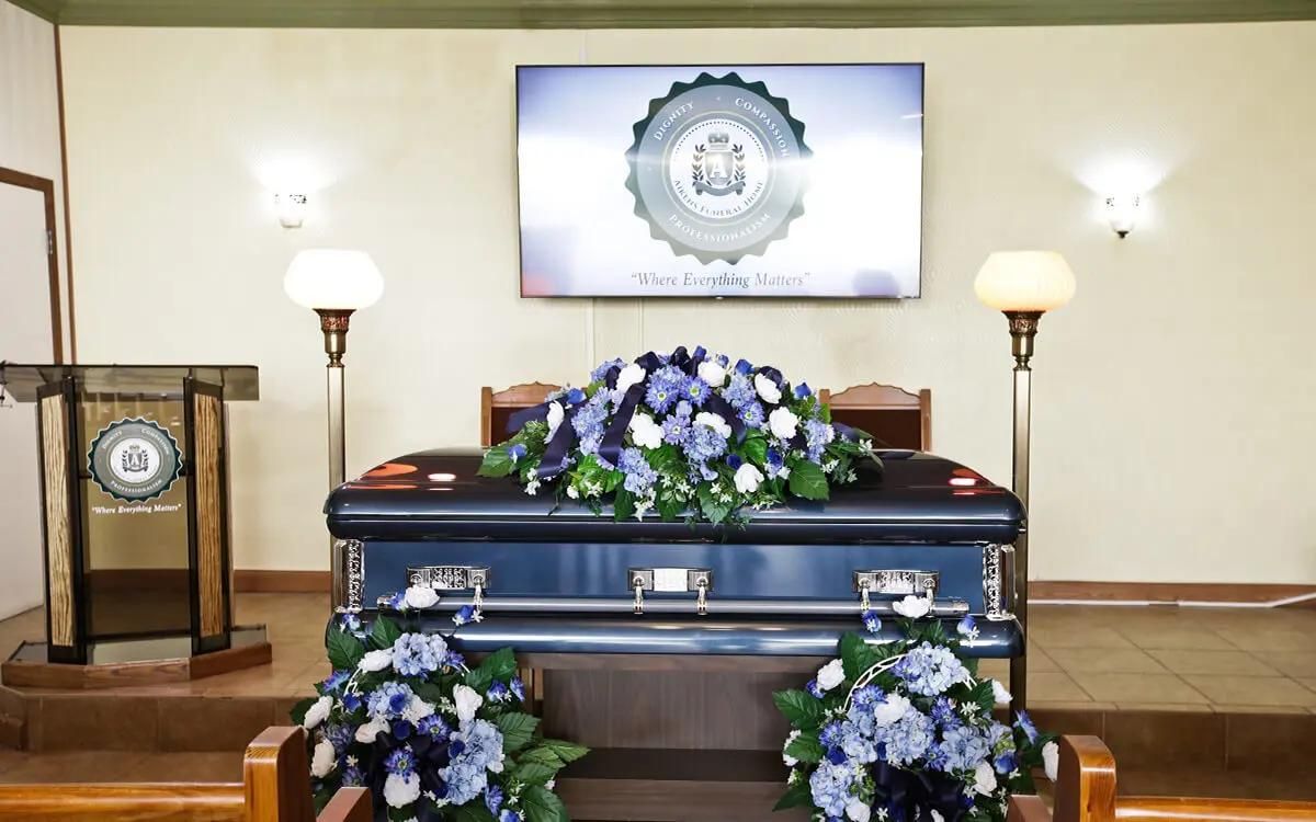 A blue casket with floral arrangements in a chapel, with a screen and lectern.