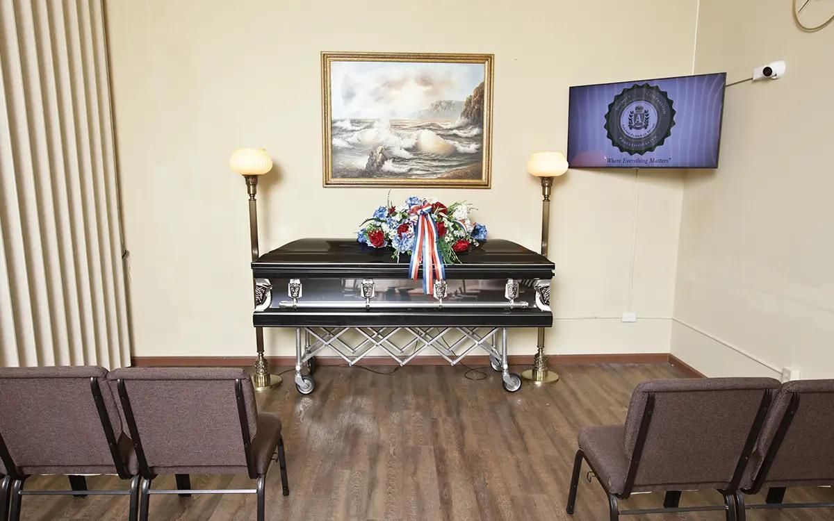 Funeral home interior: casket with floral arrangement, chairs, artwork, TV screen, lamps.
