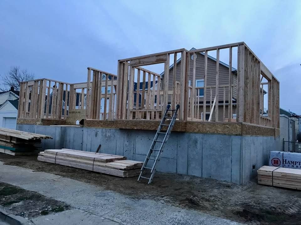 Wood frame house under construction on concrete foundation, with stacked lumber and ladder.