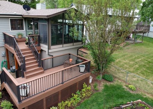 Multi-level deck with stairs, black railing, and screened-in porch next to a house with a green lawn and trees.