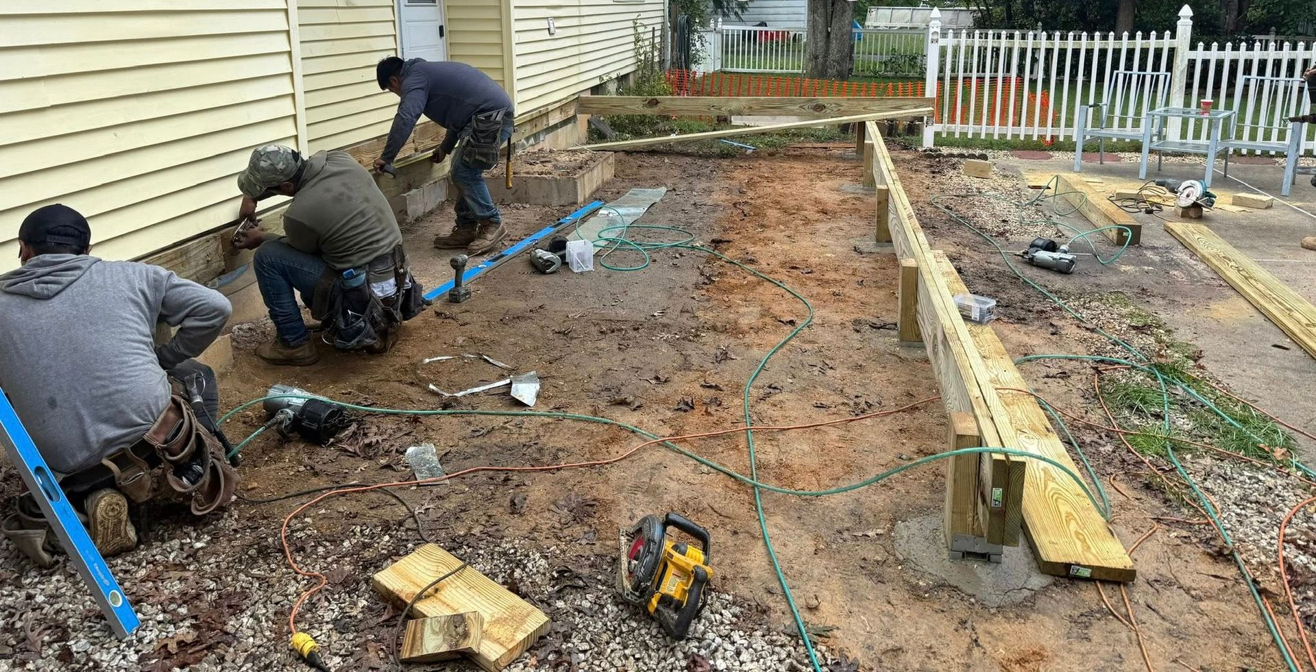Construction workers building a wooden deck near a house, using tools on a dirt ground.