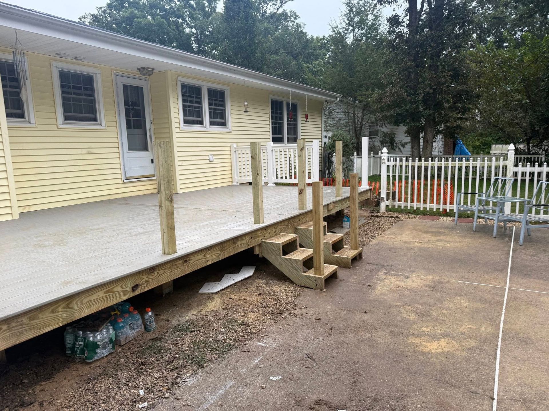 Yellow house with new wooden deck, steps, and white railing. Dirt and a white fence are in the yard.