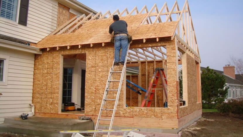 Man on a ladder constructing a wood-framed house addition; plywood siding, exposed rafters.