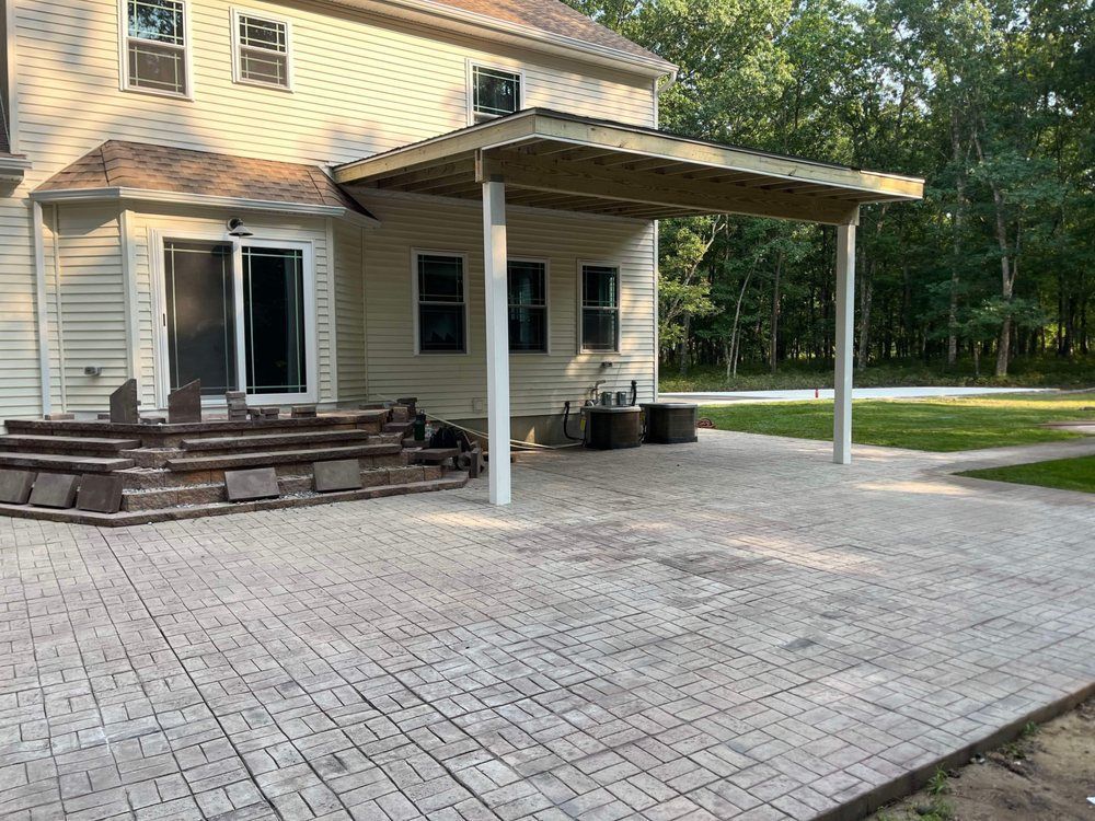 Beige house with patio and covered seating area; gray brick patio; trees in background.