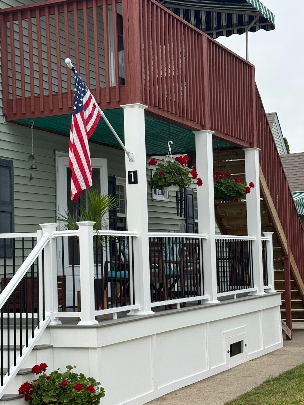 Two-story building with white porch, US flag, hanging flowers, and wooden stairs.