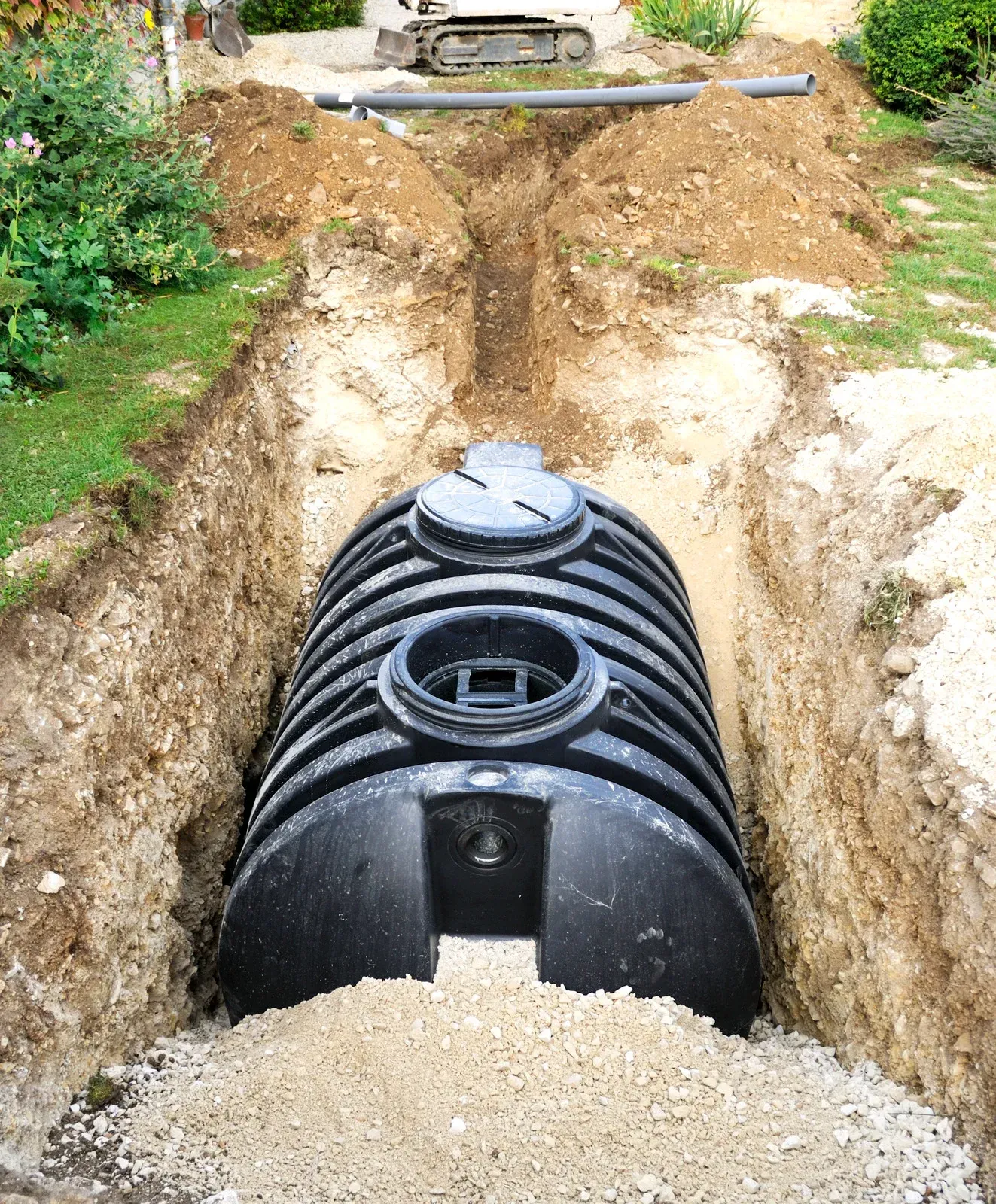 Black plastic septic tank being installed in an excavated trench, with backhoe in background.