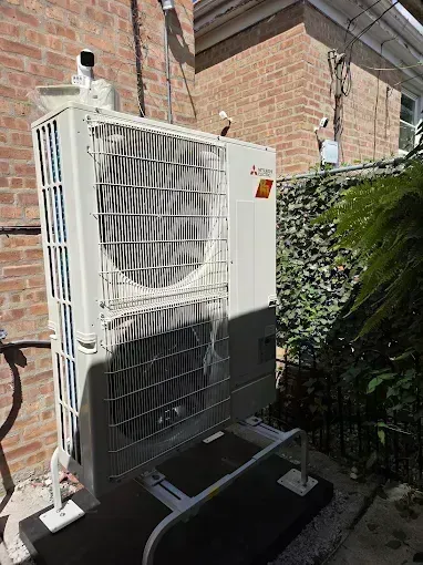 Outdoor air conditioning unit against a brick wall, with a metal safety cage, electrical lines, and a small green garden.