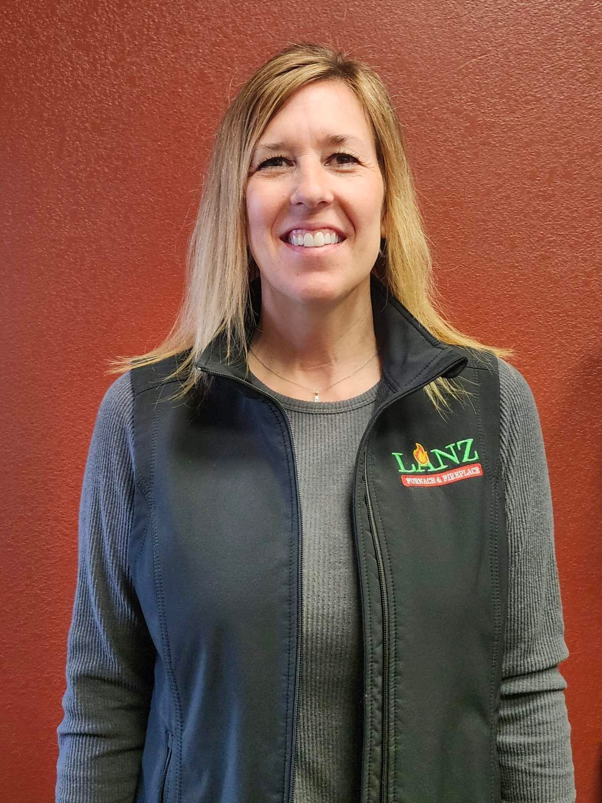 A woman wearing a black vest and a grey sweater is smiling.