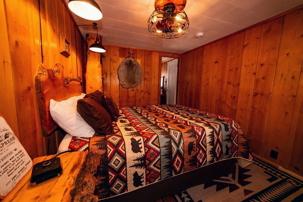 A bedroom with a bed , nightstand , lamp and wooden walls.