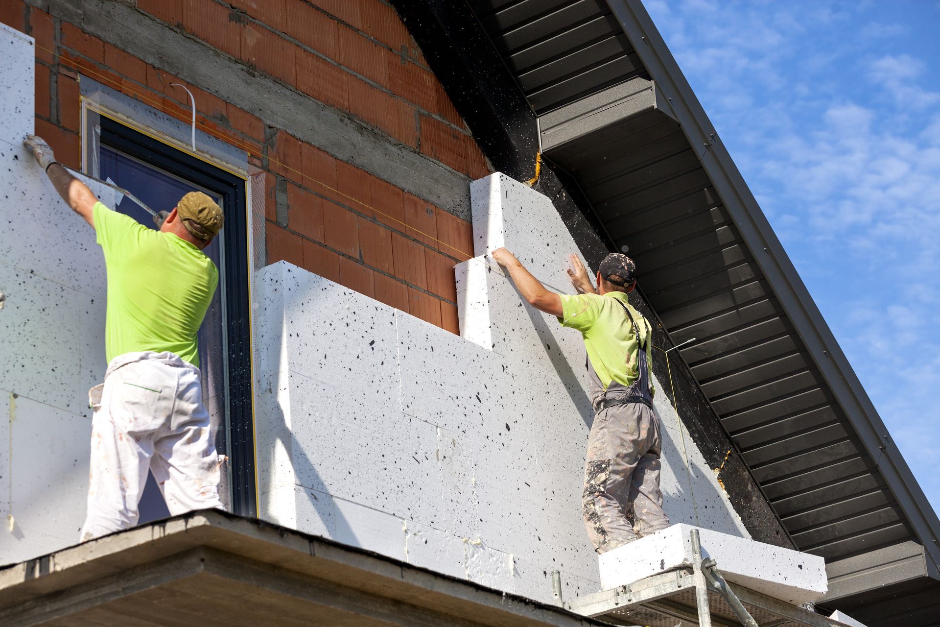 Two workers installing exterior wall insulation on a brick building under a blue sky