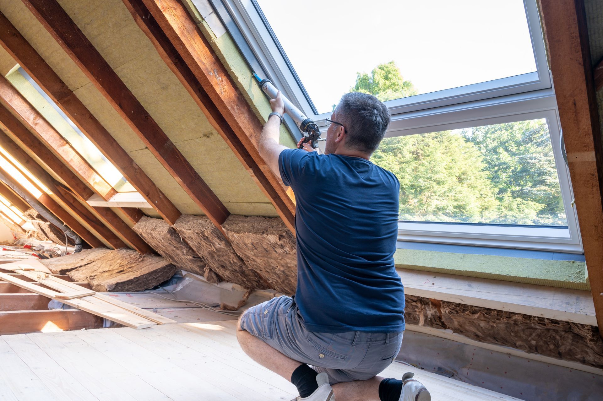 Worker kneeling in an attic, securing insulation under a slanted roof beside a window.