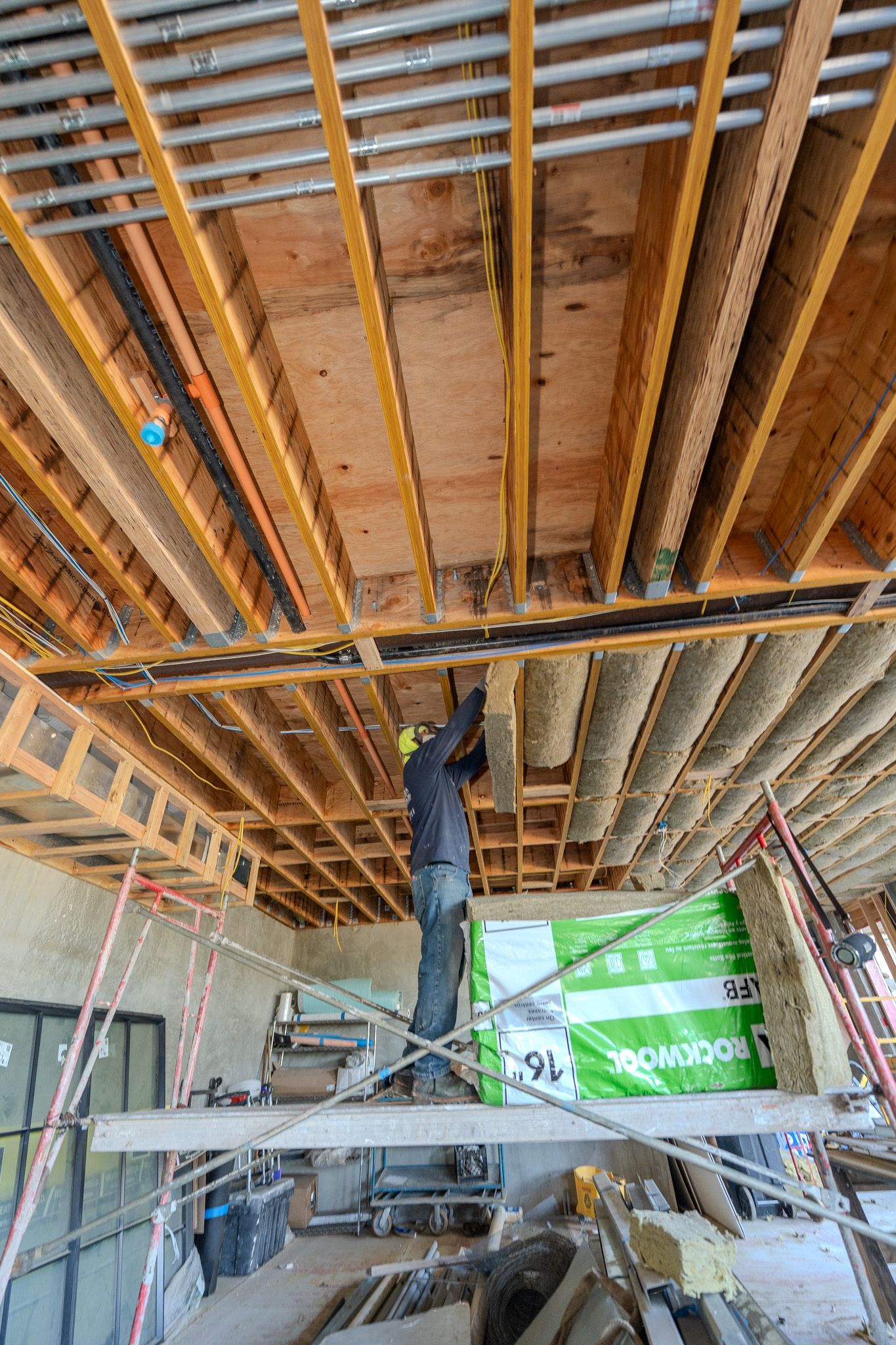 Worker installing yellow insulation in an attic, reaching up under wooden beams.