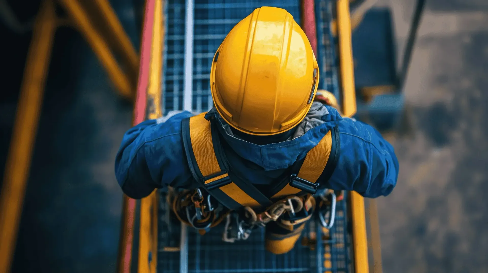 A worker in a blue jacket, yellow hard hat, and safety harness walks along a metal industrial catwalk.