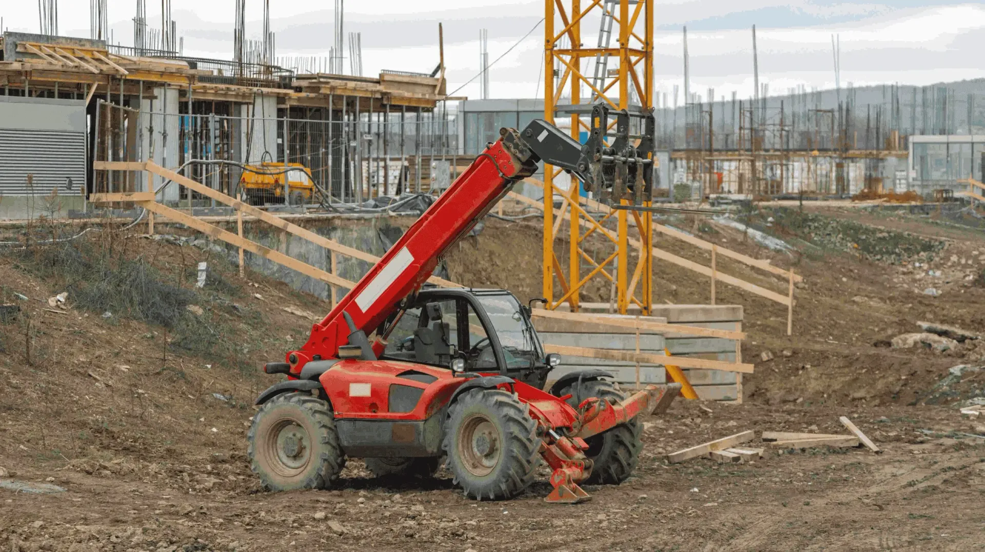 A red telescopic handler parked on a dirt construction site near a yellow tower crane and unfinished building structures.