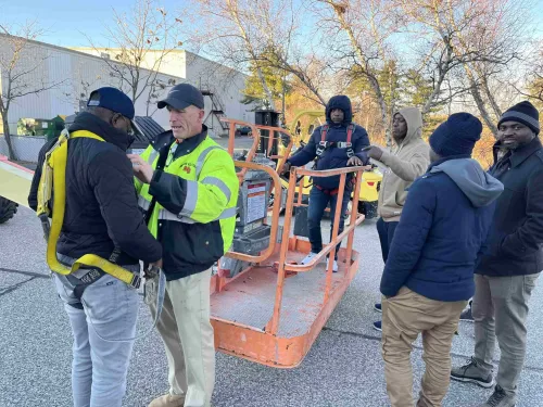 Paul Soracco of Motive Solutions LLC in a neon safety vest helps a worker put on a safety harness next to a scissor lift.