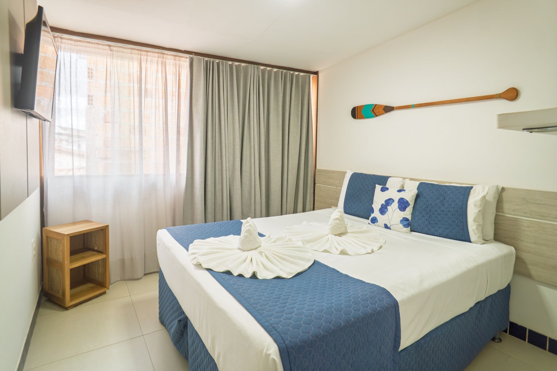 Quarto luminoso com uma cama azul e branca bem arrumada, janela com cortinas e uma pequena mesa de cabeceira de madeira.