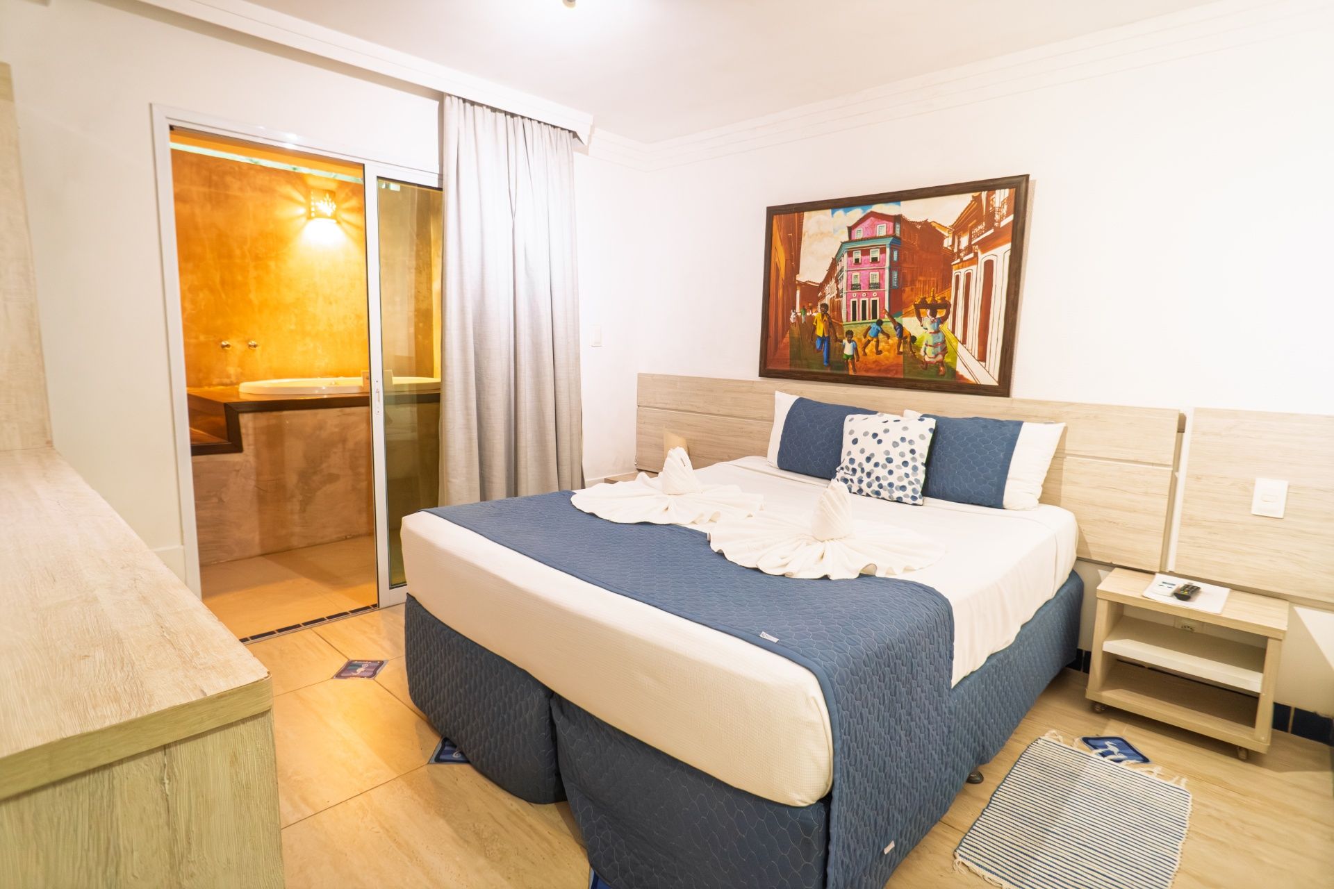 Quarto moderno e luminoso com cama de casal, detalhes em azul, mesa de cabeceira e porta aberta para o banheiro.