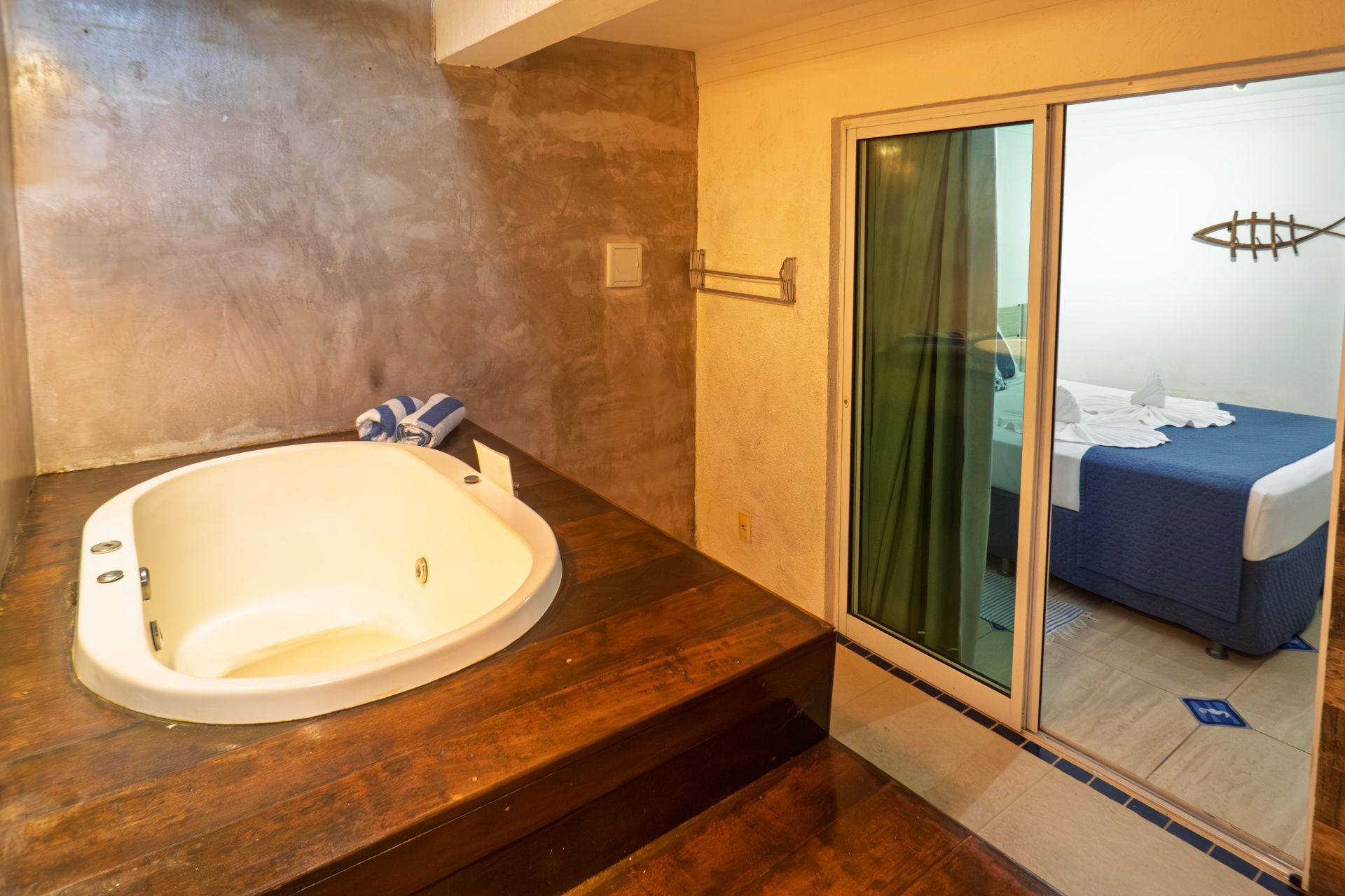 Banheiro com banheira branca ao lado de porta de correr para um quarto com cama azul e cortinas verdes.