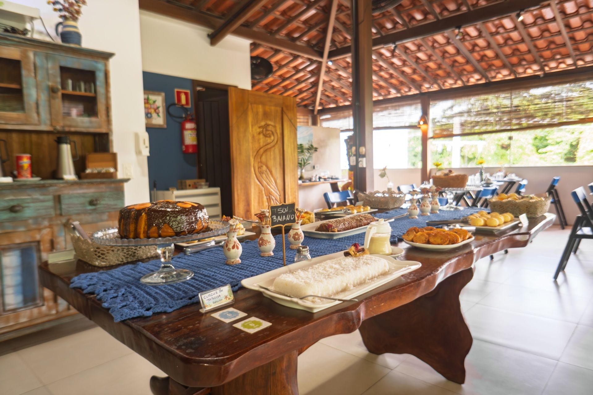 Mesa de buffet com doces e pratos em uma sala de jantar ampla e iluminada.
