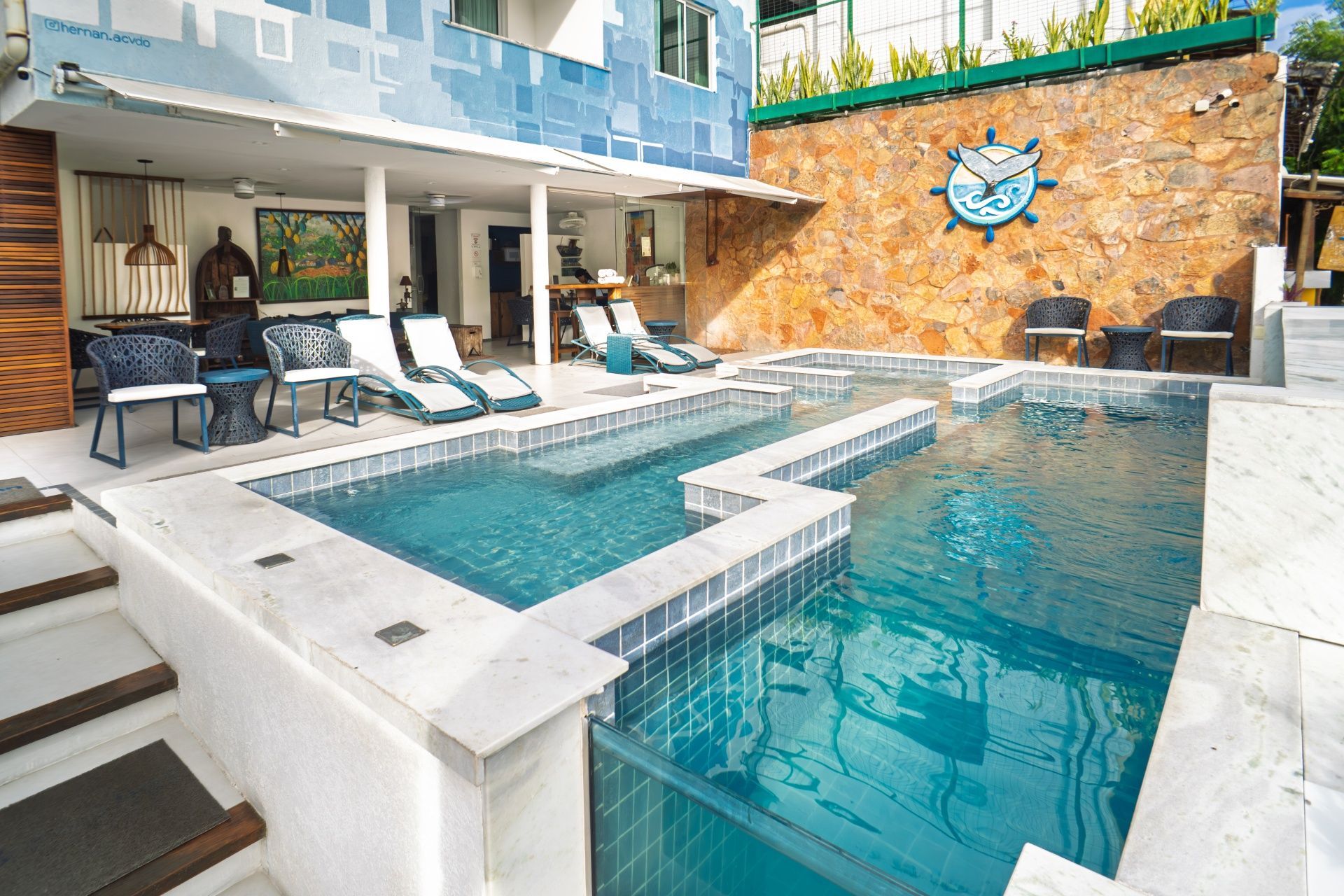 Piscina moderna no quintal com espreguiçadeiras, spa, parede de pedra e água turquesa sob um pátio coberto.