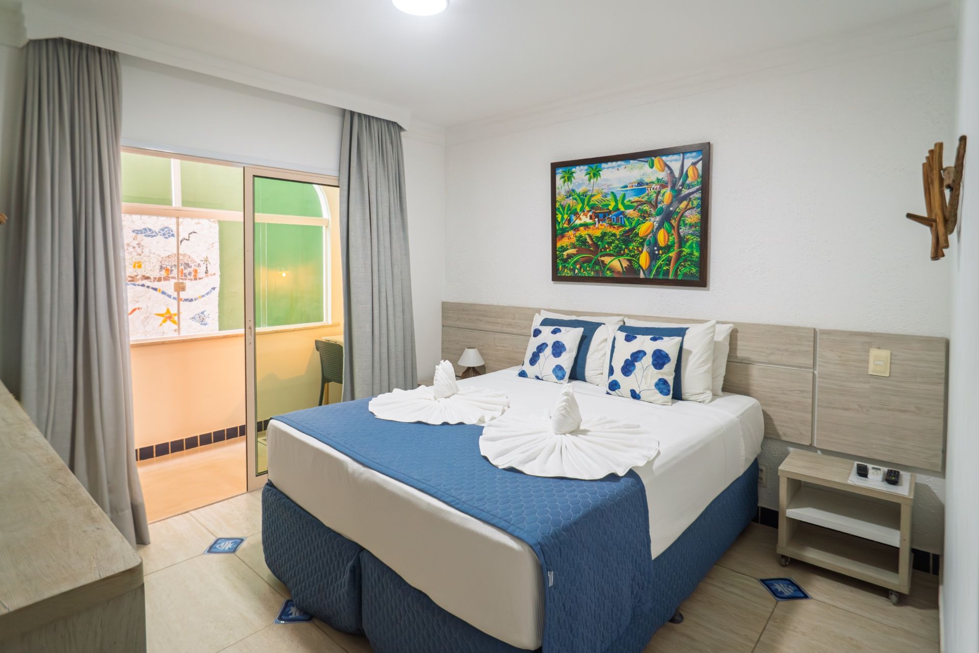 Quarto de hotel luminoso com cama bem arrumada, detalhes em azul e porta de varanda com vista para a vegetação.