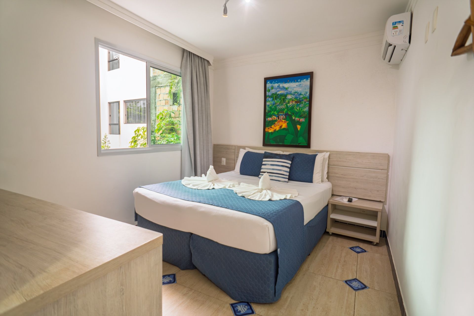 Quarto luminoso com cama arrumada, janela, obras de arte na parede e cômoda de madeira.