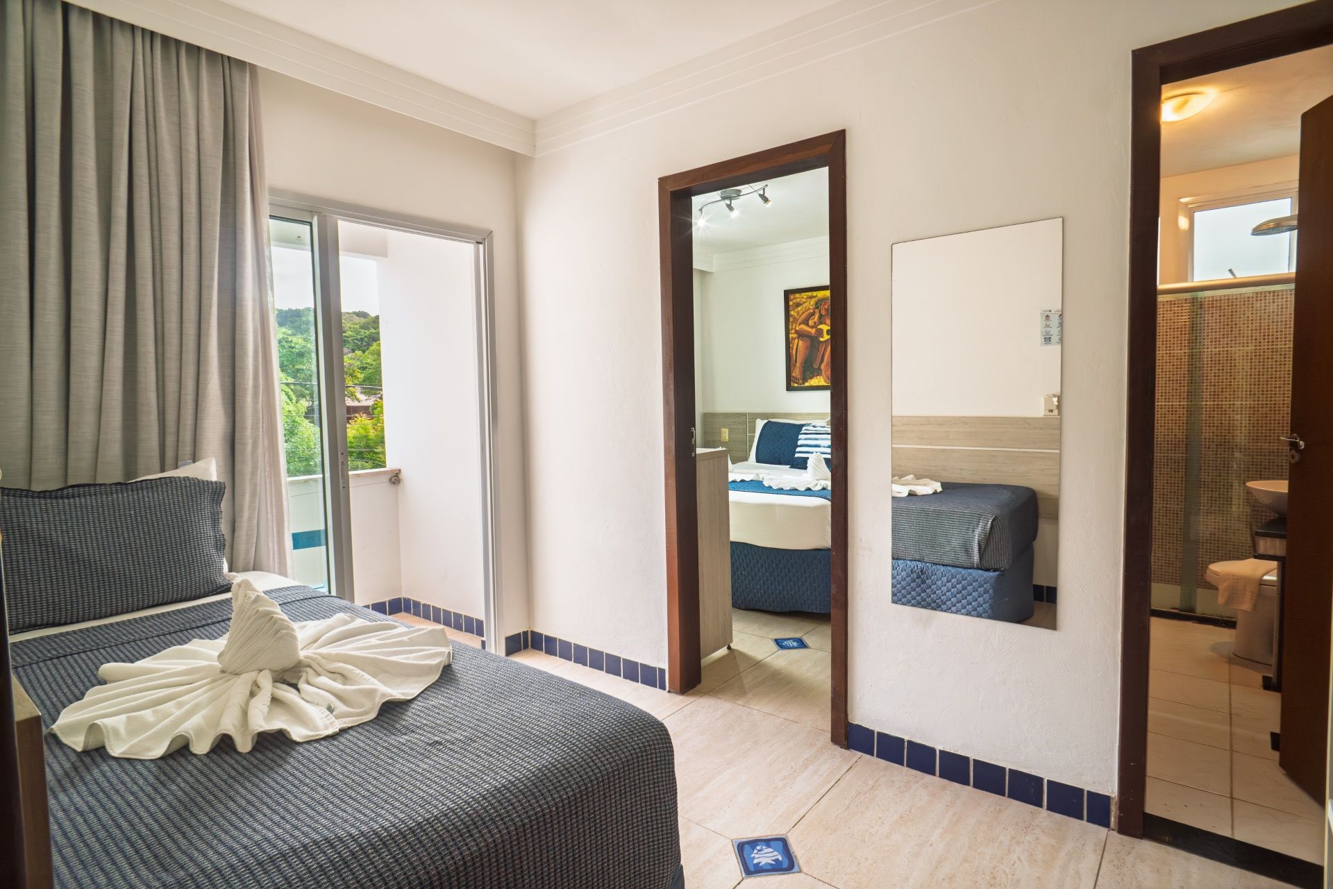 Quarto luminoso com cama, porta para varanda, espelho e porta do banheiro revestida com azulejos.