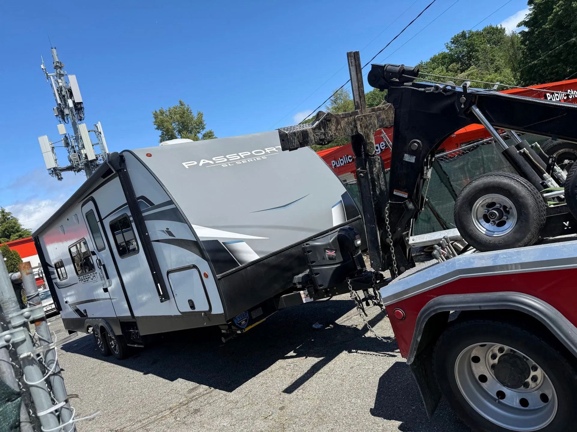 A tow truck is lifting a gray and white travel trailer next to a cell tower on a sunny day.