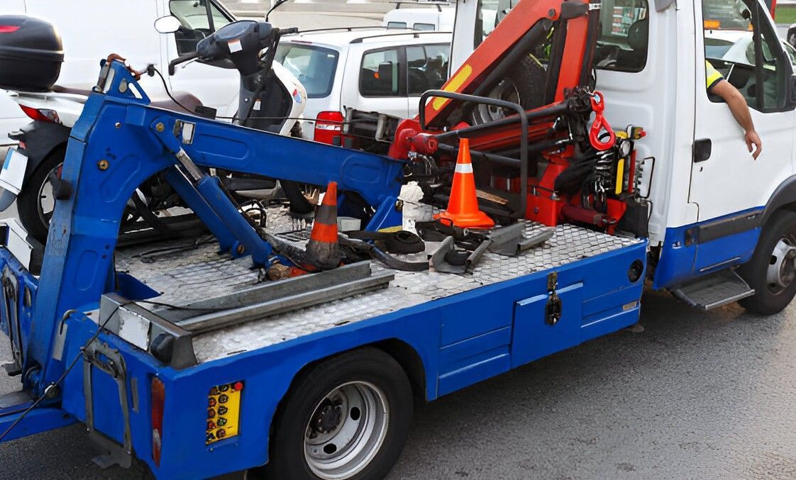 Blue and white tow truck with a crane on a flatbed; parked in a lot with a scooter and other vehicles.