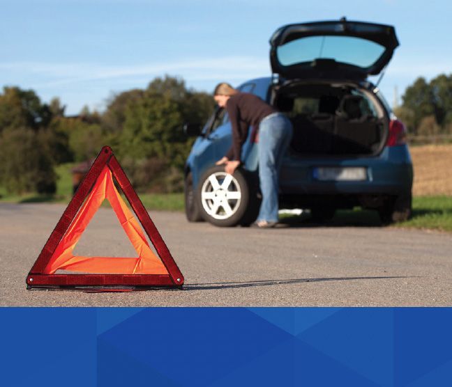 Woman changing a flat tire on the side of a road, with a safety triangle in front | McWilliams & Walden Inc.