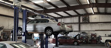 Car repair shop interior with a vehicle lifted on a hydraulic lift. A second vehicle and various tools are visible.