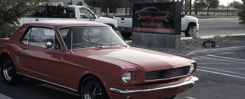 Red classic car parked in front of a sign, two trucks in the background.
