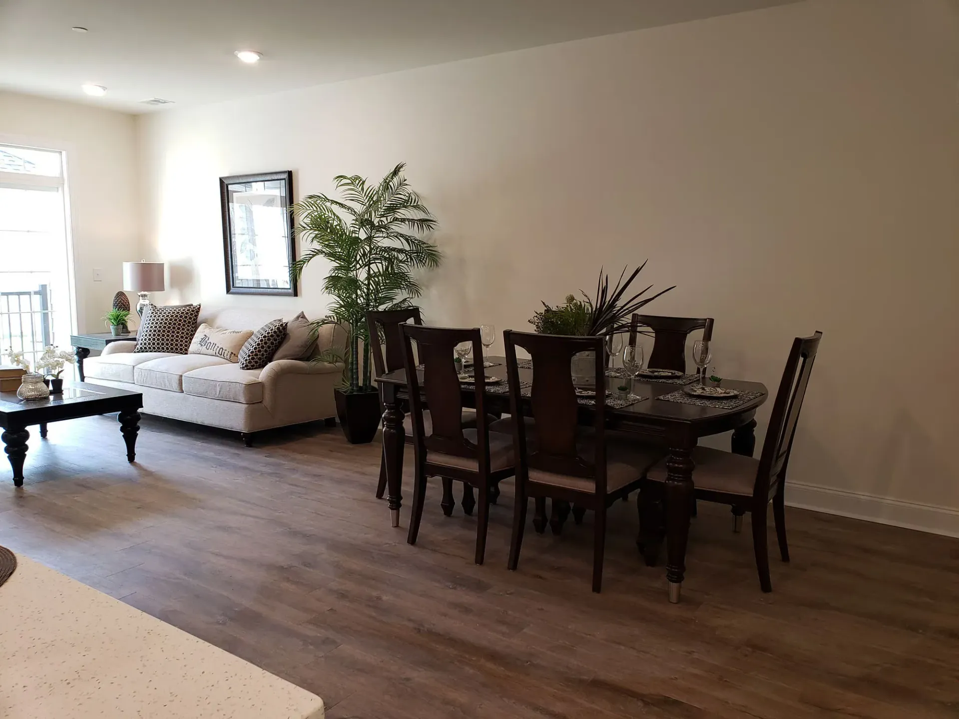 Open-concept living and dining area with a beige sofa, dark wood dining table, and potted plants at The Residences at St. Joseph's Court in Levittown, PA.