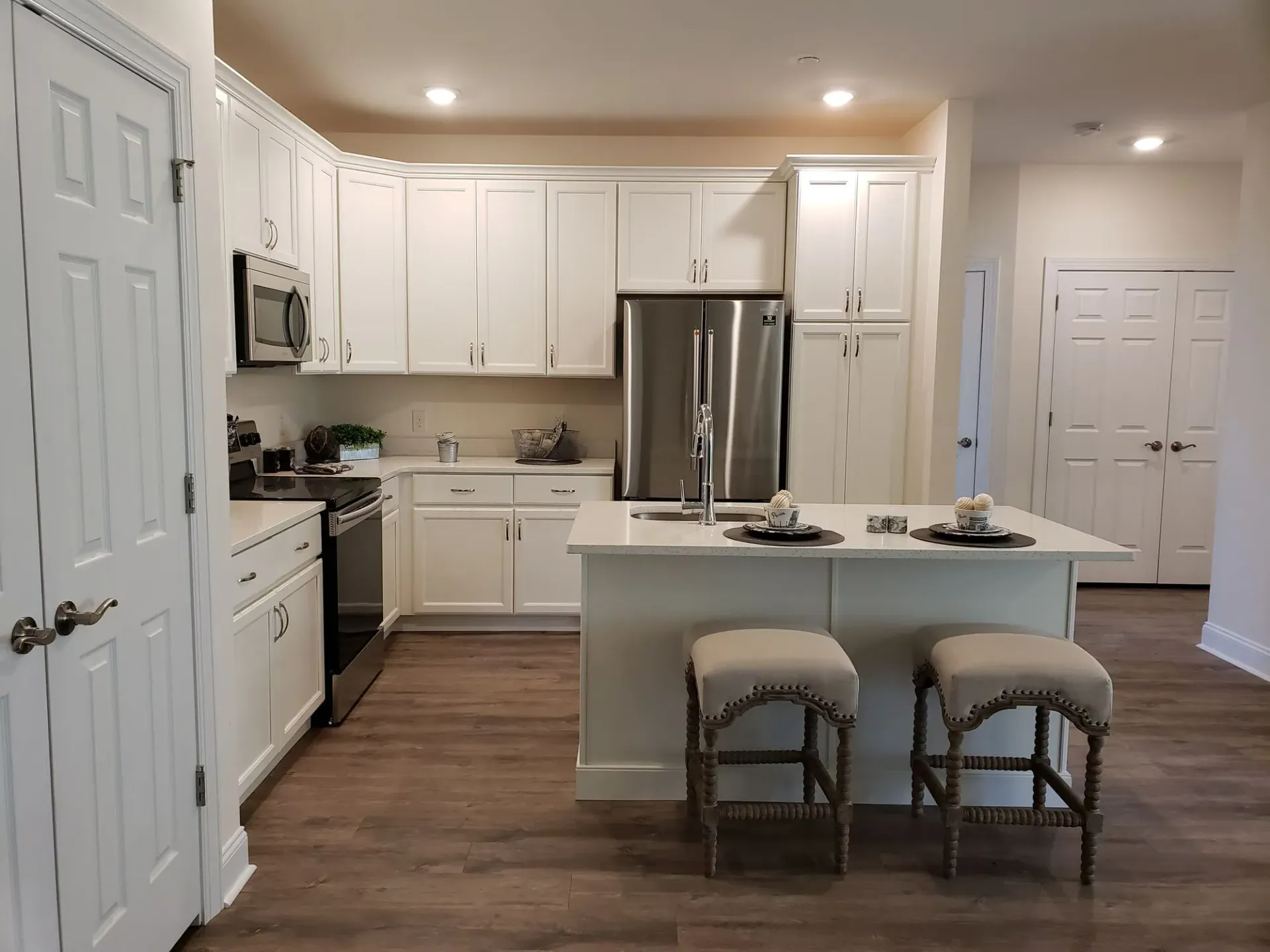 Bright white kitchen with white cabinets, stainless steel appliances, and an island with two cushioned stools at The Residences at St. Joseph's Court in Levittown, PA.