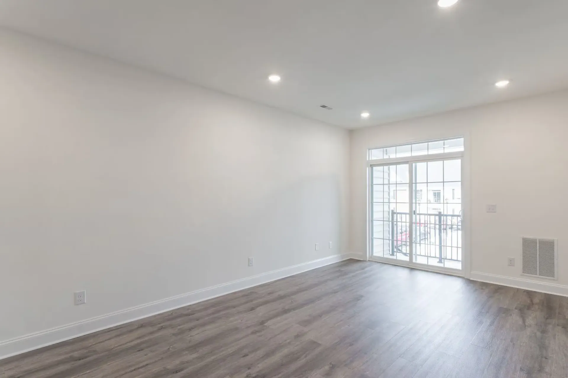 Bright empty living room with white walls, wood-look flooring, and a sliding glass door to a balcony at The Residences at St. Joseph's Court in Levittown, PA.