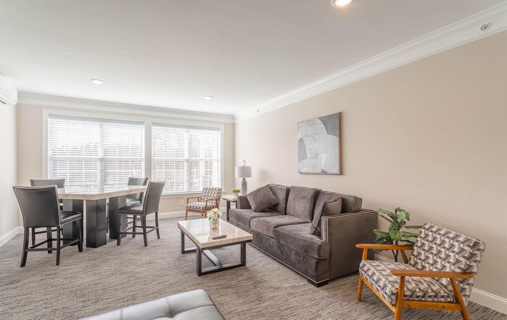 Living room and dining area in a modern apartment with beige walls, grey sofa, and large windows at The Residences at St. Joseph's Court in Levittown, PA.