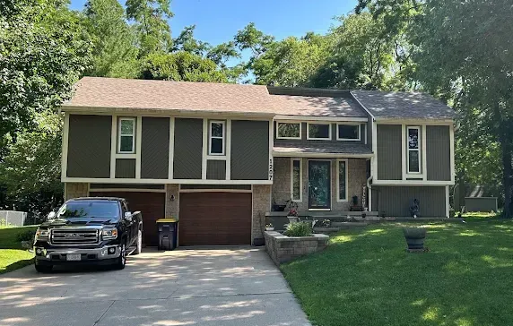 Two-story house with olive green siding, brown roof and garage doors, black truck in driveway, and green lawn.