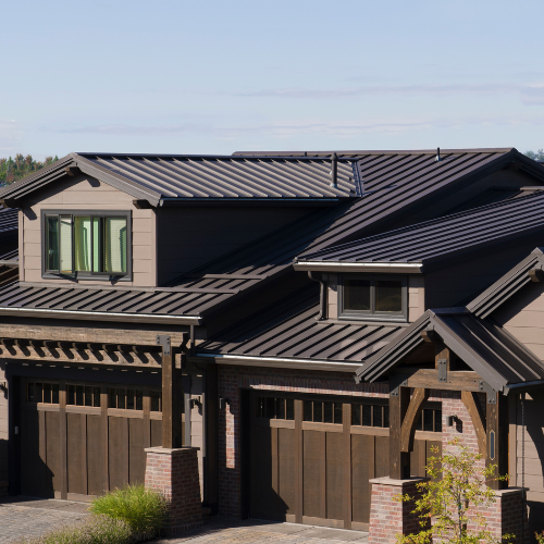 Brown two-story house with dark metal roof, wooden garage doors, and a clear blue sky.