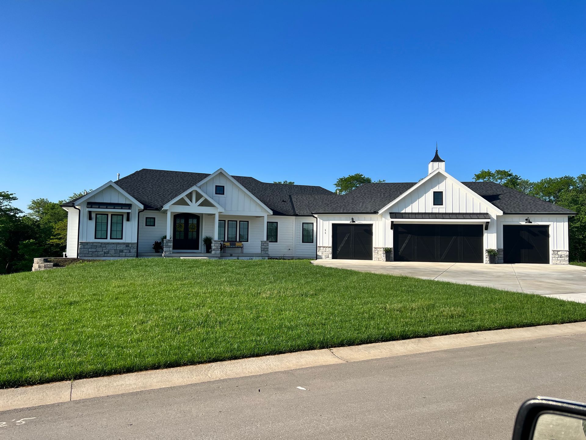 White farmhouse with black garage doors, set on a green lawn under a blue sky.