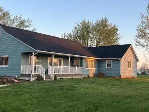 Blue ranch house with white porch and dark roof on a grassy lawn under a clear sky.
